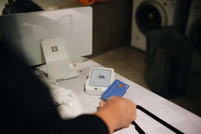 A person's hand holds a blue credit card over a white contactless payment terminal on a counter, representing a modern digital transaction crucial for customer data and AI-powered marketing.