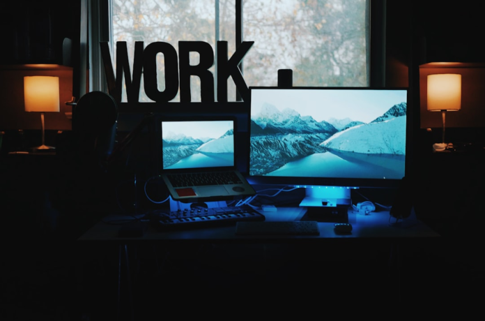 Productive home office desk featuring glowing dual monitors displaying a serene mountain landscape, a prominent WORK sign, and warm ambient lighting, representing a focused environment for digital marketing automation.