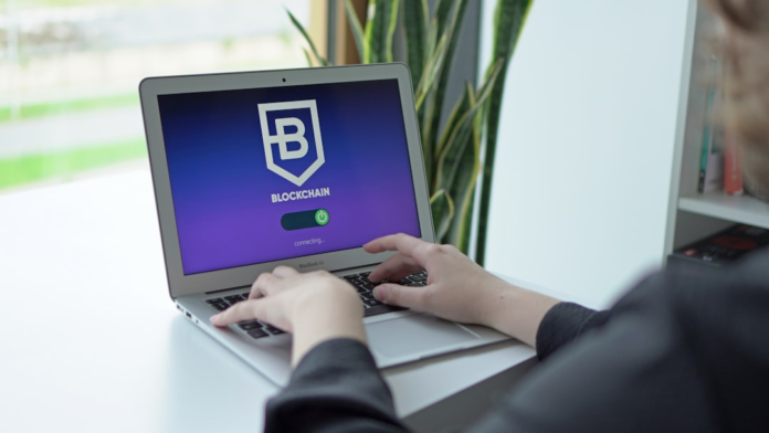 A user's hands type on a MacBook Air laptop, its screen prominently displaying a Blockchain logo with a connecting status, set within a bright, modern office.