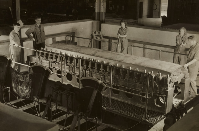 Five factory workers, men and women, observe a large, complex industrial machine in a sepia-toned vintage workshop, representing the intricate manual processes of historical business operations.