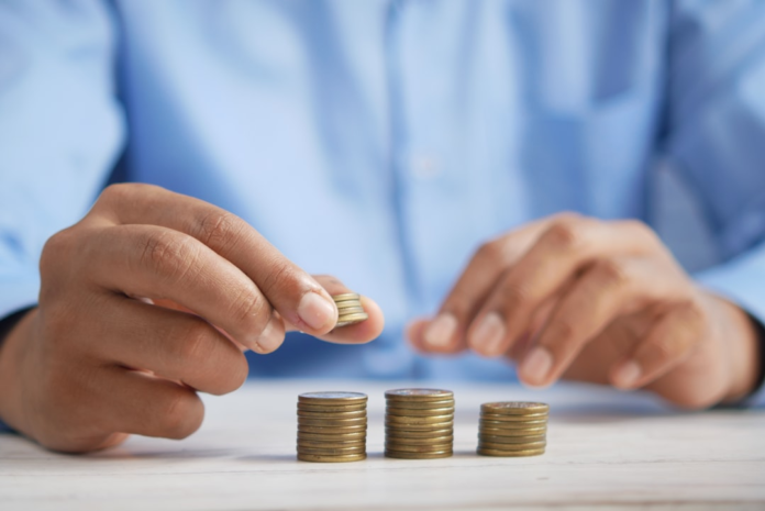 A person's hand adds a coin to one of three growing stacks on a light wooden table, symbolizing increasing earnings and financial growth from a referral program.