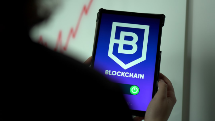 A person's hands hold a tablet displaying a vibrant BLOCKCHAIN logo, text, and toggle switch, set against a blurred background showing an upward-trending red data graph, suggesting advanced data integration and future growth in marketing.