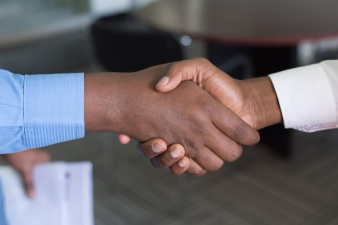 Two individuals with dark skin tones, one in a blue shirt and one in a white shirt, shake hands firmly in a professional setting, signifying trust, partnership, and successful customer interactions.