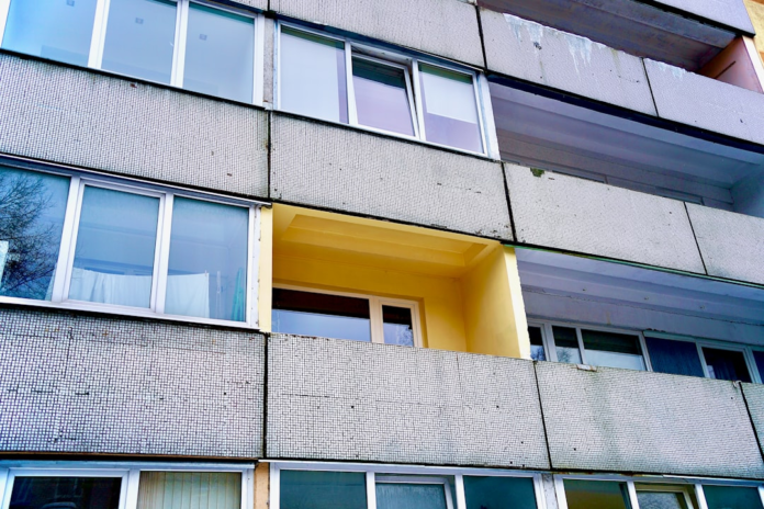 Robust urban building facade featuring textured grey panels, varied windows, and a unique bright yellow recessed balcony.