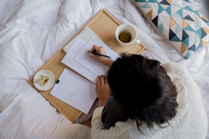 A person in a cozy bed setting uses a wooden tray to write on planner pages with a pen, embodying focused content creation for effortless Kindle ebook publishing.