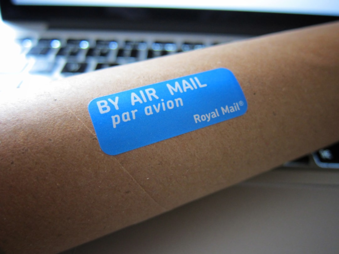 Brown mailing tube prominently displaying a blue BY AIR MAIL par avion Royal Mail sticker on a blurred laptop keyboard, illustrating the essential process of achieving successful email inbox placement.