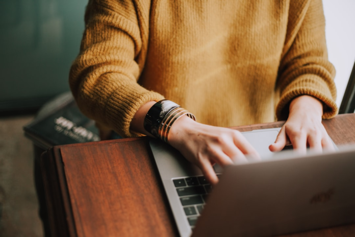 A person in a cozy mustard sweater types diligently on a laptop at a warm wooden desk, representing focused website management and the use of essential WordPress plugins.