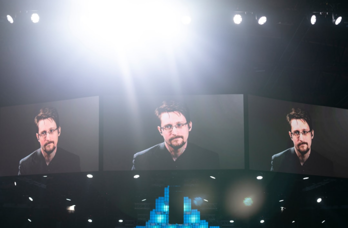 Edward Snowden's image is prominently displayed across multiple large screens in a dimly lit conference hall, highlighting the critical importance of data privacy and robust security for HIPAA compliant CRM solutions in healthcare.