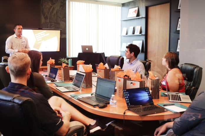 A presenter explains email marketing strategies on a screen to engaged attendees with laptops and gift bags around a conference table in a modern office.
