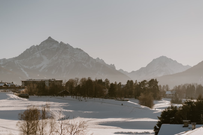 Peaceful winter landscape featuring expansive, sunlit snow-covered fields and distant buildings nestled among trees, framed by towering, snow-dusted mountain ranges under a soft, hazy sky.