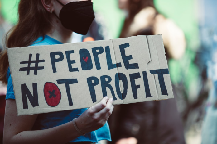 A person wearing a black mask holds a cardboard protest sign that reads '#PEOPLE NOT PROFIT', highlighting a focus on values within discussions of content monetization.