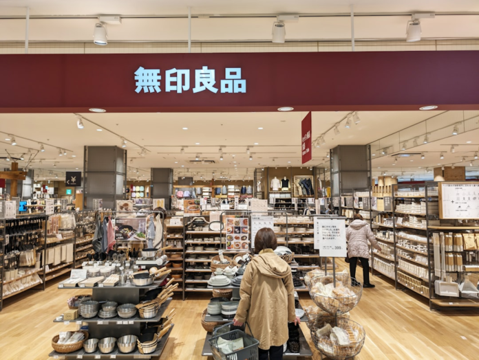 Interior of a brightly lit Muji retail store, identified by its distinctive red sign, displaying an organized array of minimalist home goods and kitchenware while customers browse products.