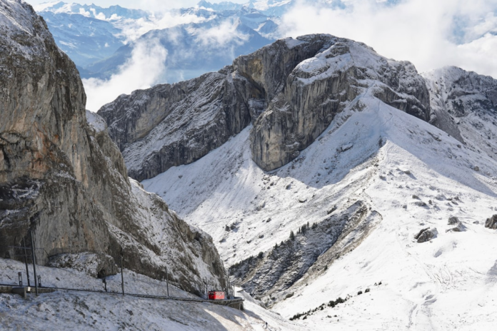 Vast snowy mountains and jagged rock faces dominate the landscape, with a small red train navigating a winding railway track through the steep terrain under a bright, cloudy sky.