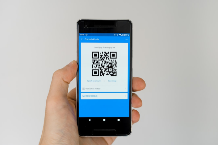 A hand holds a smartphone displaying an Alipay QR code payment screen, representing digital transaction methods relevant to Shopify merchant fees.