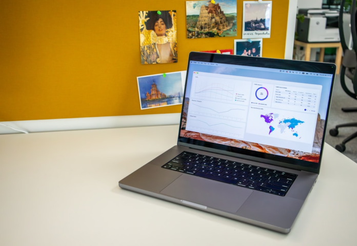 A modern laptop on a white desk displays a comprehensive web analytics dashboard featuring traffic graphs and a world map, set against a warm yellow pinboard in a professional office environment, illustrating essential SEO metrics.