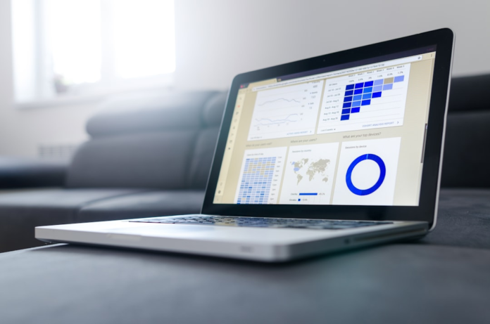Laptop screen displaying marketing analytics dashboards with charts, a world map for user location, and device usage statistics, placed on a dark sofa in a brightly lit room.